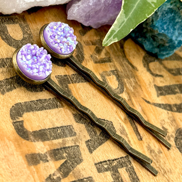 All Up In The Hair | Online Accessory Boutique Located in Mooresville, NC | Side view of two Lepidolite Druzy Bobby Pins laying on a wood background with black lettering. There are crystals and ivy leaves at the top of the image.