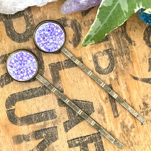 All Up In The Hair | Online Accessory Boutique Located in Mooresville, NC | Two purple druzy bobby pins are laying diagonally on a grey background, surrounded by colorful glitter.