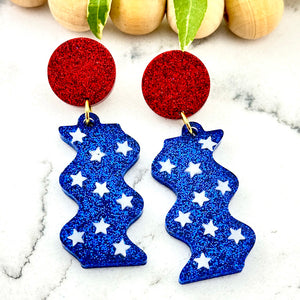 All Up In The Hair | Online Accessory Boutique Located in Mooresville, NC | Two Let Freedom Ring Earrings, featuring a red circle stud and a drop made with dark blue glitter and white stars. The earrings are laying on a white marble background. Behind the earrings is a wood bead garland and ivy leaves.