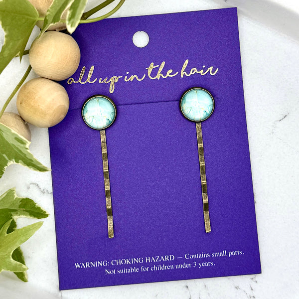 All Up In The Hair | Online Accessory Boutique Located in Mooresville, NC | Two Light Marble Bobby Pins on an indigo colored, All Up In The Hair branded packaging card. The card is laying on a white marble background, next to a wood bead garland and ivy leaves.