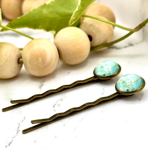 All Up In The Hair | Online Accessory Boutique Located in Mooresville, NC | Side view of two Light Marble Bobby Pins laying on a white marble background next to a wood bead garland and ivy leaves.