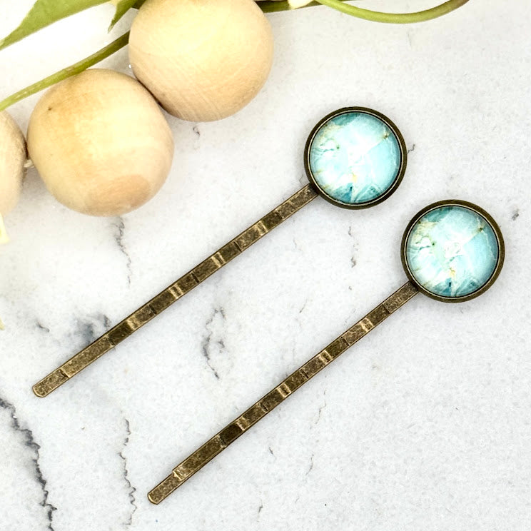 All Up In The Hair | Online Accessory Boutique Located in Mooresville, NC | Two Light Blue Marble Style Bobby Pins laying on a white marble background. The bobby pins are laying next to a wood bead garland and ivy leaves.