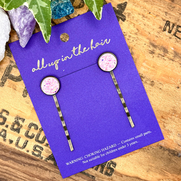 All Up In The Hair | Online Accessory Boutique Located in Mooresville, NC | Two Light Pink Druzy Bobby Pins on an indigo colored, All Up In The Hair branded packaging card. The card is laying on a wood background with black lettering. There are crystals and ivy at the top of the image.