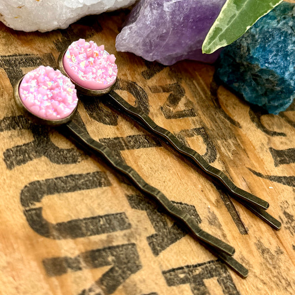 All Up In The Hair | Online Accessory Boutique Located in Mooresville, NC | Side view of two Light PInk Druzy Bobby Pins laying on a wood background with black lettering. There are crystals and ivy leaves at the top of the image.