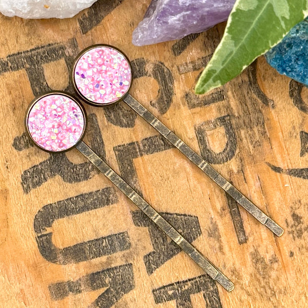 All Up In The Hair | Online Accessory Boutique Located in Mooresville, NC | Two Light Pink Druzy Bobby Pins laying diagonally on a wood background with black lettering. There are crystals and ivy at the top of the image.
