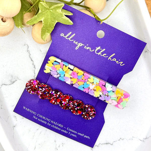 All Up In The Hair | Online Accessory Boutique Located in Mooresville, NC | Our Love Struck Barrette Set on an indigo colored, All Up In The Hair branded packaging card. The card is laying on a white marble background. At the top of the image is a wood bead garland and ivy leaves.