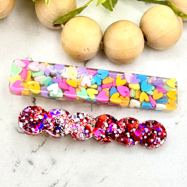 All Up In The Hair | Online Accessory Boutique Located in Mooresville, NC | A rectangle barrette filled with pastel hearts and a red, purple, and silver glitter barrette laying on a white marble background. At the top of the image is a wood bead garland and ivy leaves.