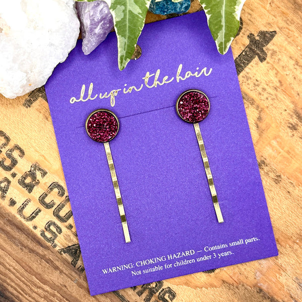 All Up In The Hair | Online Accessory Boutique Located in Mooresville, NC | An All Up In The Hair branded packaging card is laying on a grey background, surrounded by colorful glitter. On the card is two magenta druzy bobby pins.