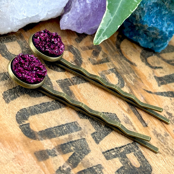 All Up In The Hair | Online Accessory Boutique Located in Mooresville, NC | Side view of two Magenta Druzy Bobby Pins on a wood background with black lettering. There are crystals and ivy leaves at the top of the image.