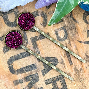 All Up In The Hair | Online Accessory Boutique Located in Mooresville, NC | Two magenta druzy bobby pins are laying diagonally on a grey background, surrounded by colorful glitter.