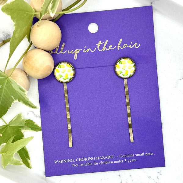 All Up In The Hair | Online Accessory Boutique Located in Mooresville, NC | Two Make Lemonade Bobby Pins on an indigo colored, All Up In The Hair branded packaging card. The card is laying on a white marble background with a wood bead garland and ivy leaves.