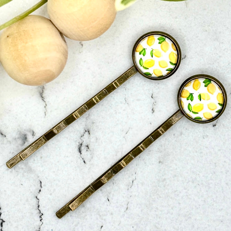 All Up In The Hair | Online Accessory Boutique Located in Mooresville, NC | Two Lemon Bobby Pins on a white marble background next to a wood bead garland and ivy leaves.