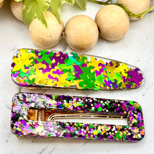 All Up In The Hair | Online Accessory Boutique Located in Mooresville, NC | Two barrettes in Mardi Gras colors laying on a grey background, surrounded by confetti hearts,