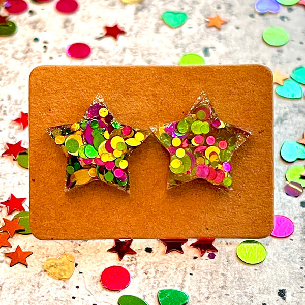 All Up In The Hair | Online Accessory Boutique Located in Mooresville, NC | Two purple, green, and gold star earrings on a small kraft card on a grey background. The card is surrounded by colorful confetti.