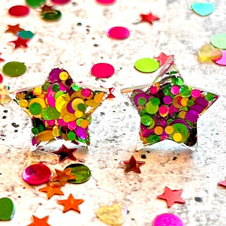 All Up In The Hair | Online Accessory Boutique Located in Mooresville, NC | Two purple, green, and gold glitter star earrings laying on a grey background, surrounded by colorful glitter.