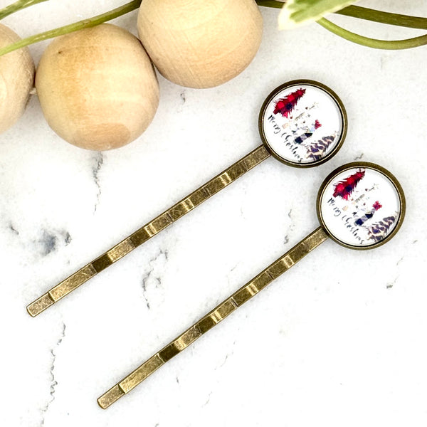 All Up In The Hair | Online Accessory Boutique Located in Mooresville, NC | Two Merry Christmas Bobby Pins laying on a white marble background with a wood bead garland and ivy leaves.