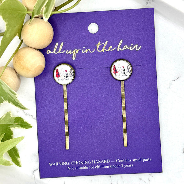 All Up In The Hair | Online Accessory Boutique Located in Mooresville, NC | Two Merry Christmas Bobby Pins on an indigo colored, All Up In The Hair branded packaging card. The card is laying on a gray background, surrounded by colorful glitter.