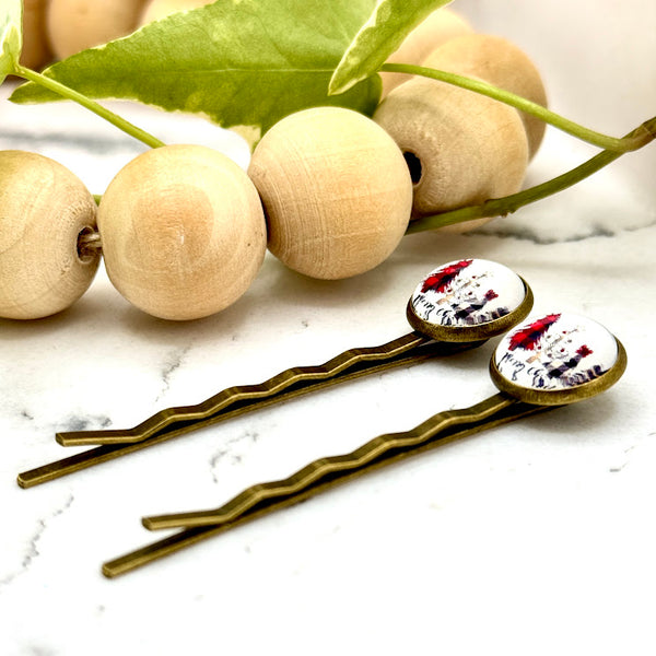 All Up In The Hair | Online Accessory Boutique Located in Mooresville, NC | Side view of two Merry Christmas Bobby Pins on a white marble background with a wood bead garland and ivy leaves.