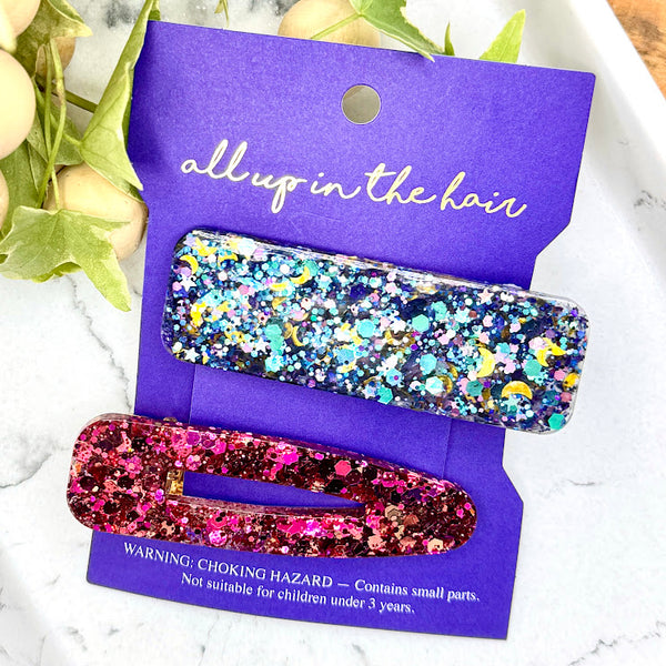 All Up In The Hair | Online Accessory Boutique Located in Mooresville, NC | An All Up In The Hair branded indigo colored backer card is laying on a grey background surrounded by silver star glitter. Attached to the card is a blue/purple rectangular barrette and a pink glitter teardrop barrette.