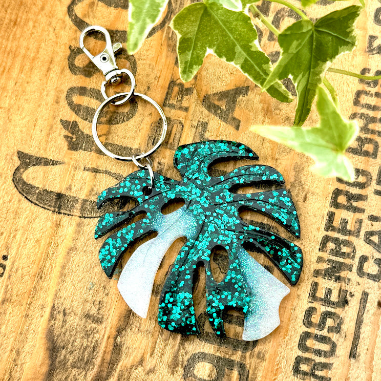 All Up In The Hair | Online Accessory Boutique Located in Mooresville, NC | Resin Monstera Albo Leaf Key Chain on a wood background with black lettering.  There are ivy leaves next to the keychain.