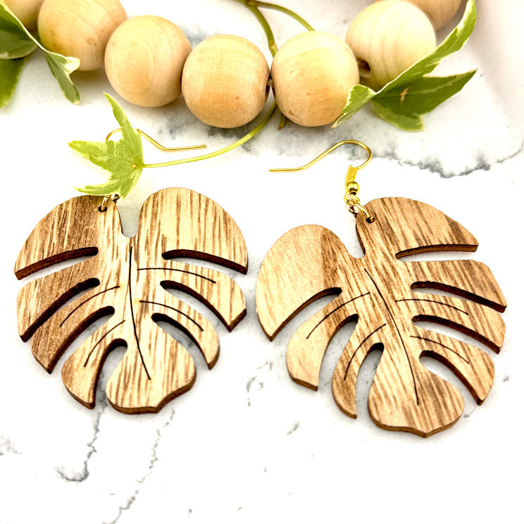 All Up In The Hair | Online Accessory Boutique Located in Mooresville, NC | Two Wood  Monstera Leaf Earrings on a white marble background. Behind the earrings is a wood bead garland and ivy leaves.