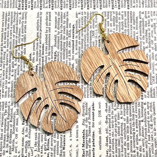 All Up In The Hair | Online Accessory Boutique Located in Mooresville, NC | Two Wood Monstera Earrings laying on a book page.