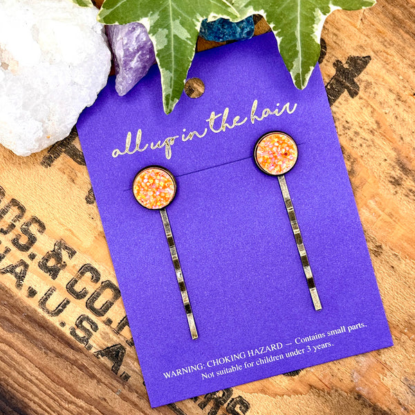 All Up In The Hair | Online Accessory Boutique Located in Mooresville, NC | Two Neon Orange Druzy Bobby Pins on an indigo colored, All Up In The Hair branded packaging card. The card is laying on a wood background with black lettering. There are crystals and ivy leaves at the top of the image.