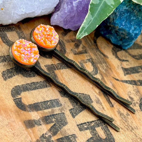 All Up In The Hair | Online Accessory Boutique Located in Mooresville, NC | Side view of two Neon Orange Druzy Bobby Pins laying on a wood background with black lettering. There are crystals and ivy leaves at the top of the image.