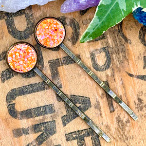 All Up In The Hair | Online Accessory Boutique Located in Mooresville, NC | Two Neon Orange Druzy Bobby Pins on a wood background with black lettering. There are crystals and ivy leaves at the top of the image.