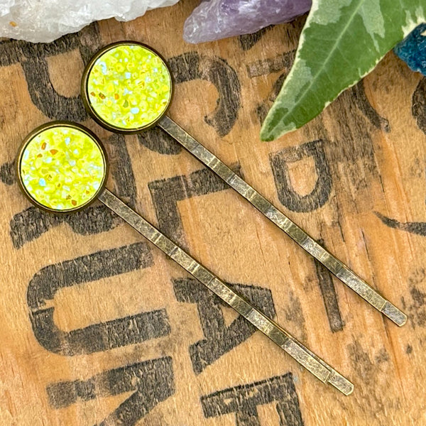 All Up In The Hair | Online Accessory Boutique Located in Mooresville, NC | Two neon yellow druzy bobby pins laying diagonally on a grey background, surrounded by colorful glitter.