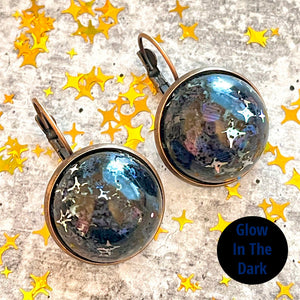 All Up In The Hair | Online Accessory Boutique Located in Mooresville, NC | Two black iridescent dangle earrings laying on a grey background, surrounded by gold star glitter. In the bottom right hand corner of the image is a black circle that says "glow in the dark" in dark blue letters.