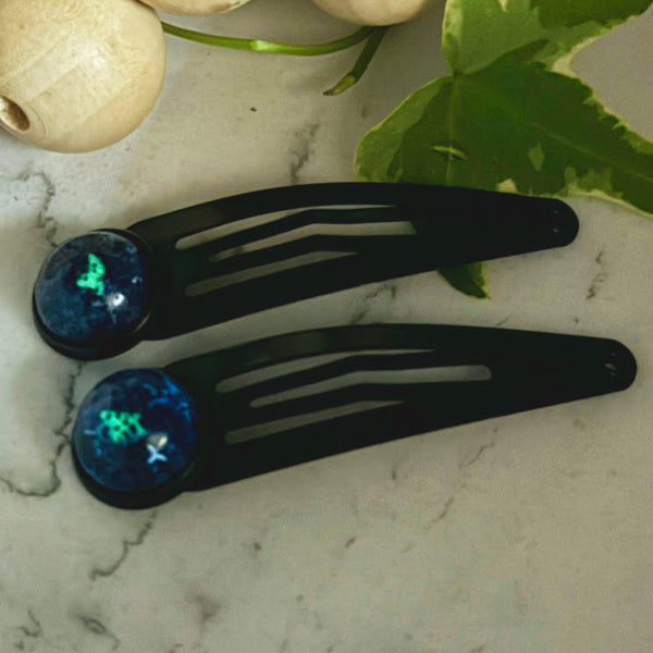 All Up In The Hair | Online Accessory Boutique Located in Mooresville, NC | Two Nova Snap Barrettes glowing dark blue and green on a white marble background. At the top of the image is a wood bead garland and ivy leaves.