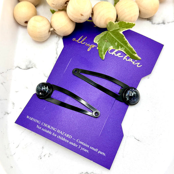 All Up In The Hair | Online Accessory Boutique Located in Mooresville, NC | Two Nova Snap Barrettes on an indigo colored, All Up In The Hair branded packaging card. The card is laying on a white marble background. At the top of the image is a wood bead garland and ivy leaves.