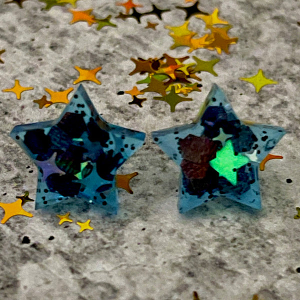All Up In The Hair | Online Accessory Boutique Located in Mooresville, NC | Two star earrings on a grey background surrounded by gold star glitter. The stars are glowing a light blue and the white stars inside them are glowing green.