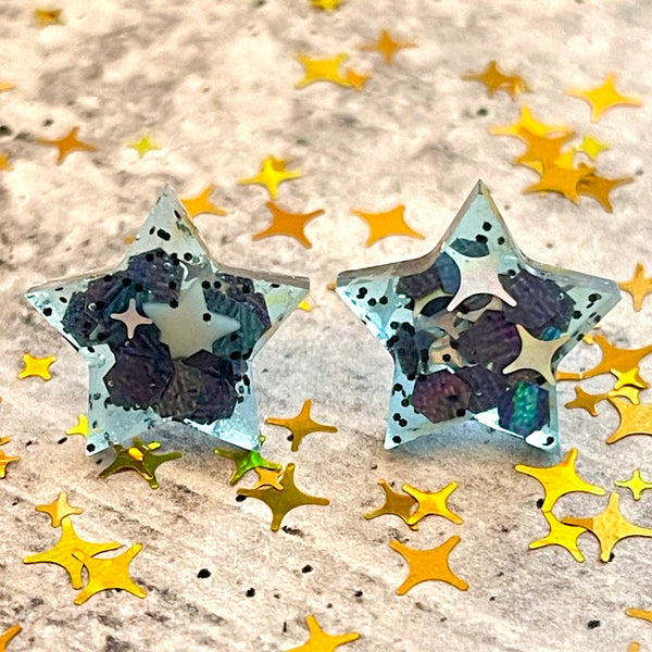 All Up In The Hair | Online Accessory Boutique Located in Mooresville, NC | Two star earrings on a grey background surrounded by gold star glitter. The stars are a clear blue, with iridescent black glitter and white and silver stars.