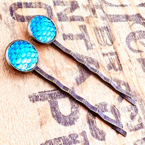 All Up In The Hair | Online Accessory Boutique Located in Mooresville, NC | Side view of two Ocean Blue Mermaid Bobby Pins laying on a wood background with black lettering.