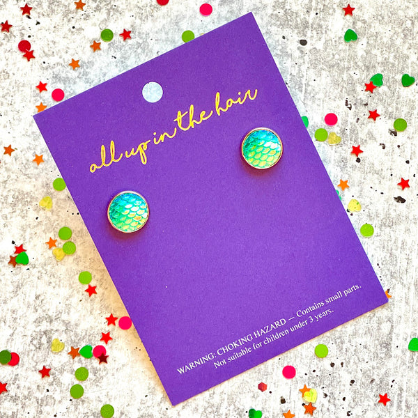 All Up In The Hair | Online Accessory Boutique Located in Mooresville, NC | Two ocean blue mermaid earrings on an indigo colored, All Up In The Hair branded packaging card. The card is laying on a gray background, surrounded by colorful glitter.