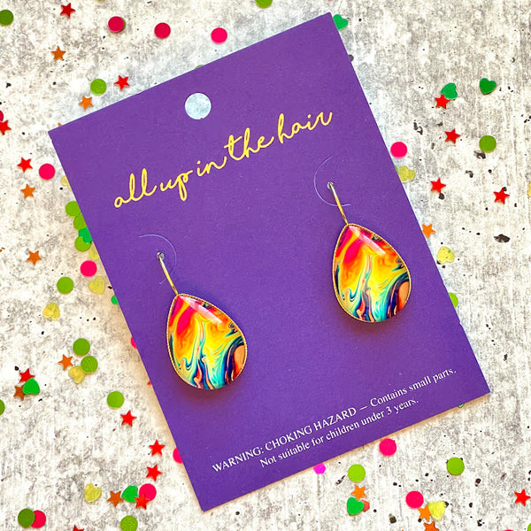 All Up In The Hair | Online Accessory Boutique Located in Mooresville, NC | Two teardrop oil slick earrings on an indigo colored, All Up In The Hair branded packaging card. The card is laying on a gray background, surrounded by colorful glitter.