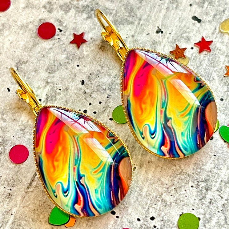 All Up In The Hair | Online Accessory Boutique Located in Mooresville, NC | Two teardrop earrings with an oil slick looking design, laying on a gray table, surrounded by colorful glitter.
