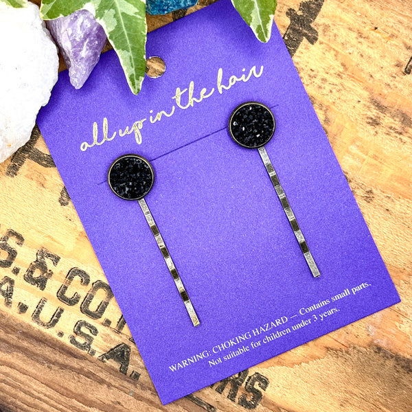 All Up In The Hair | Online Accessory Boutique Located in Mooresville, NC | Two Onyx Druzy Bobby Pins on an indigo colored, All Up In The Hair branded packaging card. The card is laying on a wood background with black lettering. There are crystals and ivy leaves at the top of the image.