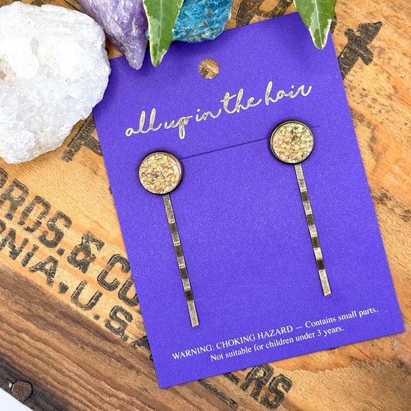 All Up In The Hair | Online Accessory Boutique Located in Mooresville, NC | Two Peach Bellini Druzy Bobby Pins on an indigo colored, All Up In The Hair branded packaging card. The card is laying on a wood background with black lettering. There are crystals and ivy leaves at the top of the image.