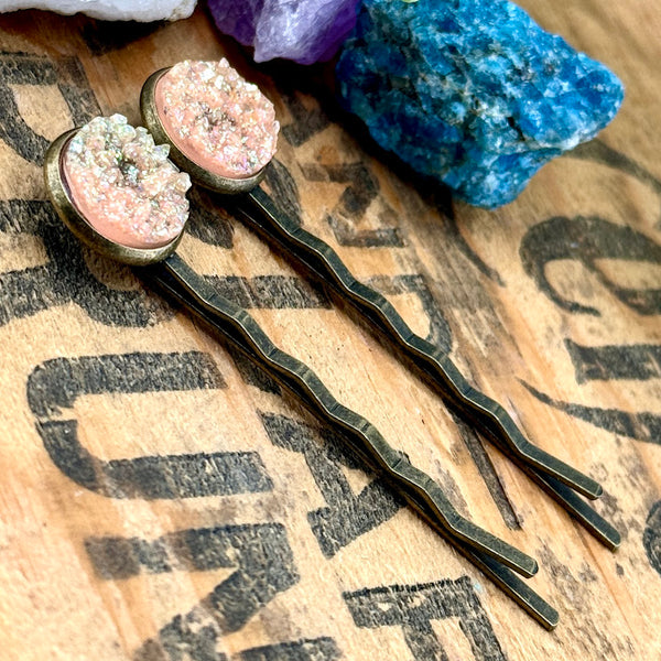 All Up In The Hair | Online Accessory Boutique Located in Mooresville, NC | Side view of our Peach Bellini Druzy Bobby Pins on a wood background with black lettering. There are crystals and ivy leaves at the top of the image.