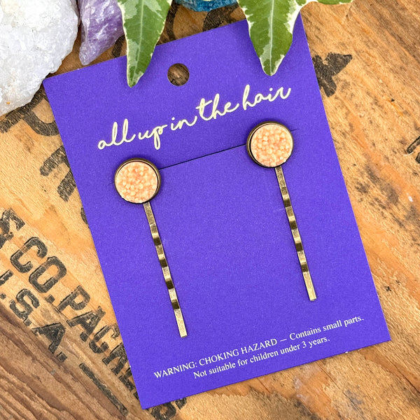 All Up In The Hair | Online Accessory Boutique Located in Mooresville, NC | Two Peach Moonstone Druzy Bobby Pins on an indigo colored, All Up In The Hair branded packaging card. The card is laying on a wood background with black lettering. There are crystals and ivy leaves at the top of the image.