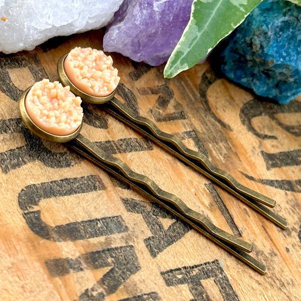 All Up In The Hair | Online Accessory Boutique Located in Mooresville, NC | Side view of two Peach Moonstone Druzy Bobby Pins laying on a wood background with black lettering. There are crystals and ivy leaves at the top of the image.