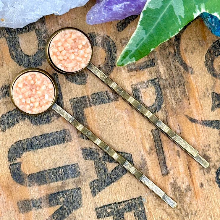 All Up In The Hair | Online Accessory Boutique Located in Mooresville, NC | Two peach moonstone druzy bobby pins laying diagonally on a grey background, surrounded by colorful glitter.
