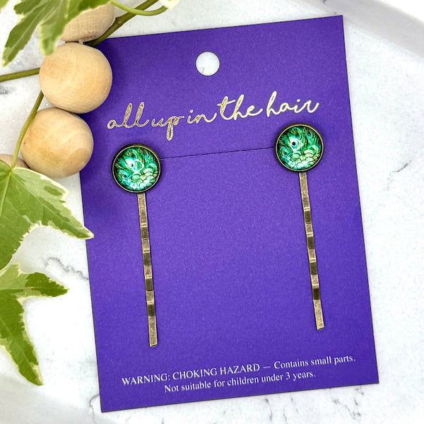 All Up In The Hair | Online Accessory Boutique Located in Mooresville, NC | Two Peacock Feather Bobby Pins on an indigo colored, All Up In The Hair branded packaging card. The card is laying on a white marble background next to a wood bead garland and ivy leaves.