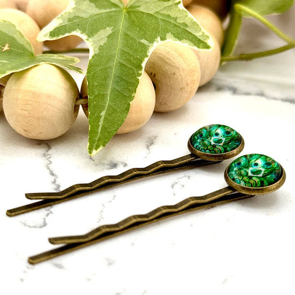 All Up In The Hair | Online Accessory Boutique Located in Mooresville, NC | Side view of two Peacock Feather Bobby Pins on a white marble background next to a wood bead garland and ivy leaves.