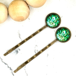 All Up In The Hair | Online Accessory Boutique Located in Mooresville, NC | Two Peacock Feather Bobby Pins laying on a white marble background next to a wood bead garland and ivy leaves.