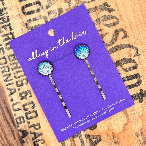 All Up In The Hair | Online Accessory Boutique Located in Mooresville, NC | Two Periwinkle Mermaid Bobby Pins on an indigo colored, All Up In The Hair branded packaging card. The card is laying on a wood background with black lettering.