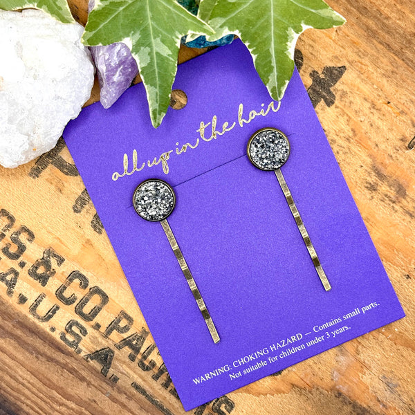 All Up In The Hair | Online Accessory Boutique Located in Mooresville, NC | Two Pewter Druzy Bobby Pins on an indigo colored, All Up In The Hair branded packaging card. The card is laying on a wood background with black lettering. There are crystals and ivy leaves at the top of the image.